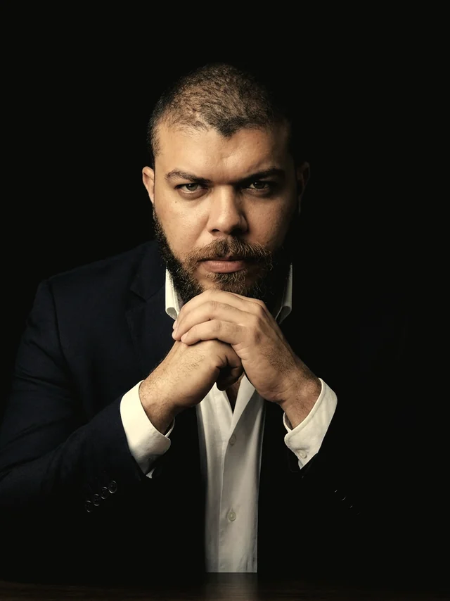 Wallison Nascimento, de terno escuro, mãos entrelaçadas sob o queixo, olhando para a câmera.